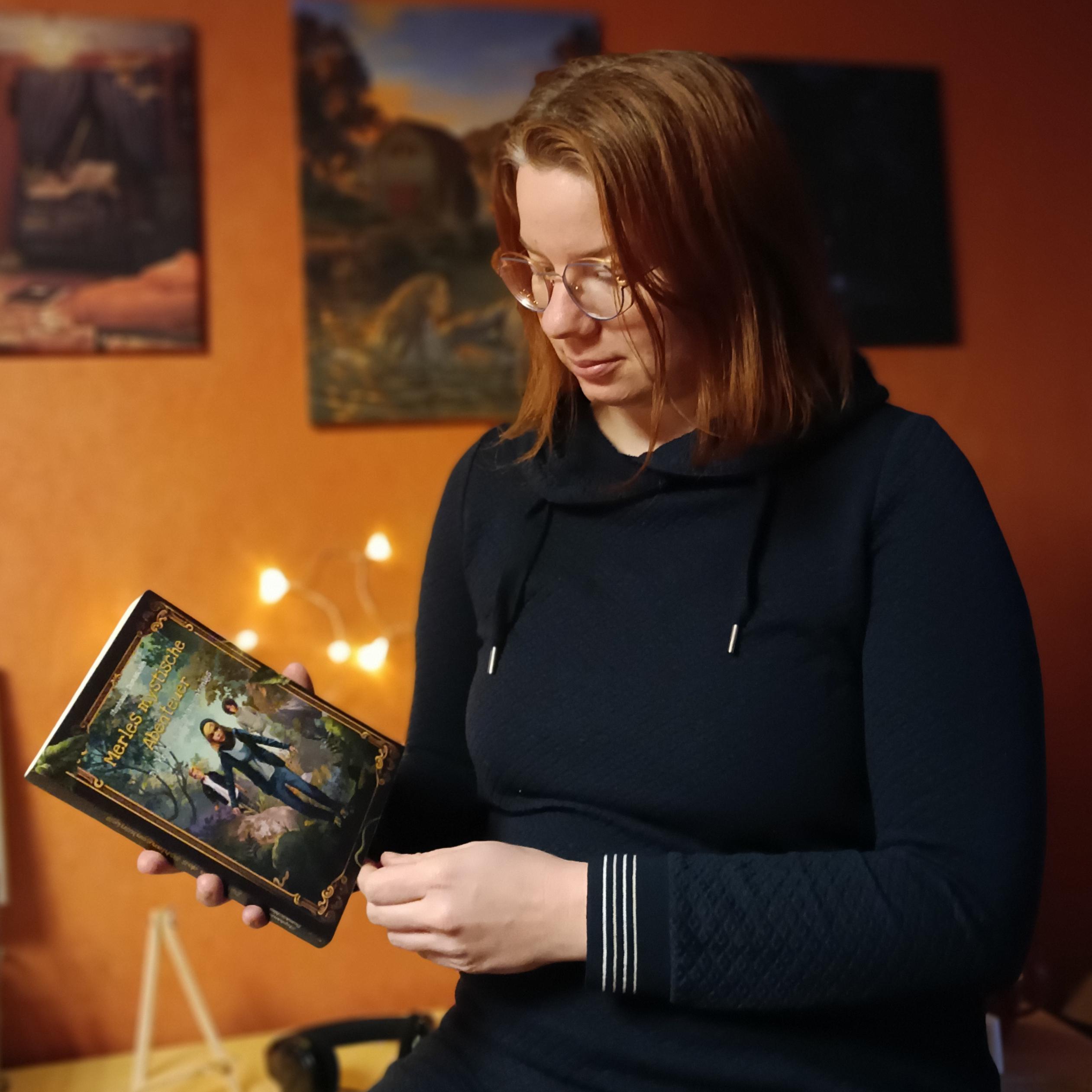 Die Autorin Stephanie Domascke hält ihr Buch Merle und die Krone des Schlangenkönigs in die Hand. Dabei steht sie vor einer Wand.