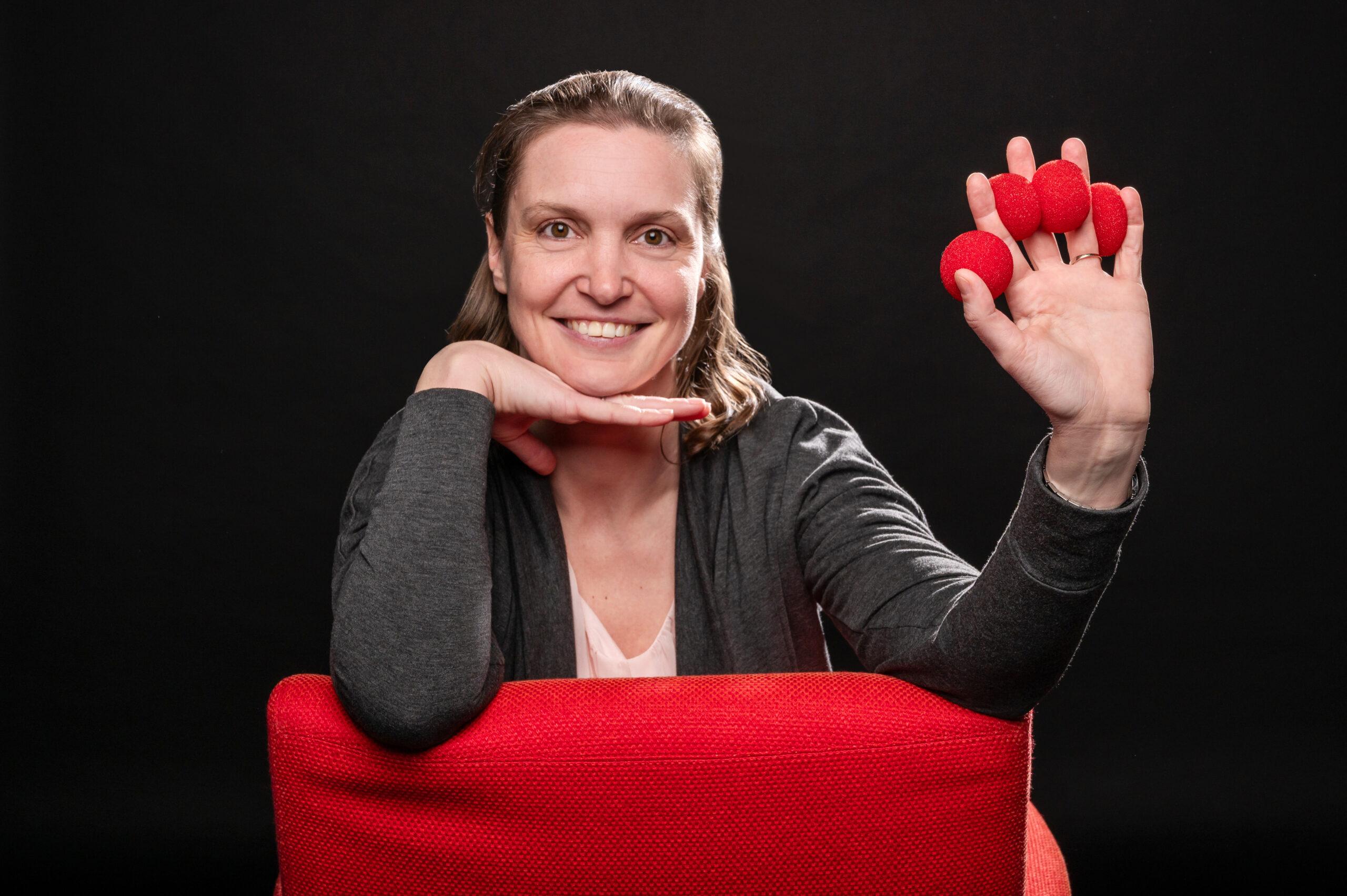 Autorin Julia Nachtigall hält Zauberzubehör in der Hand und sitzt auf einem rotem Stuhl vor schwarzem Hintergrund zum Beitrag Warum und Wieso